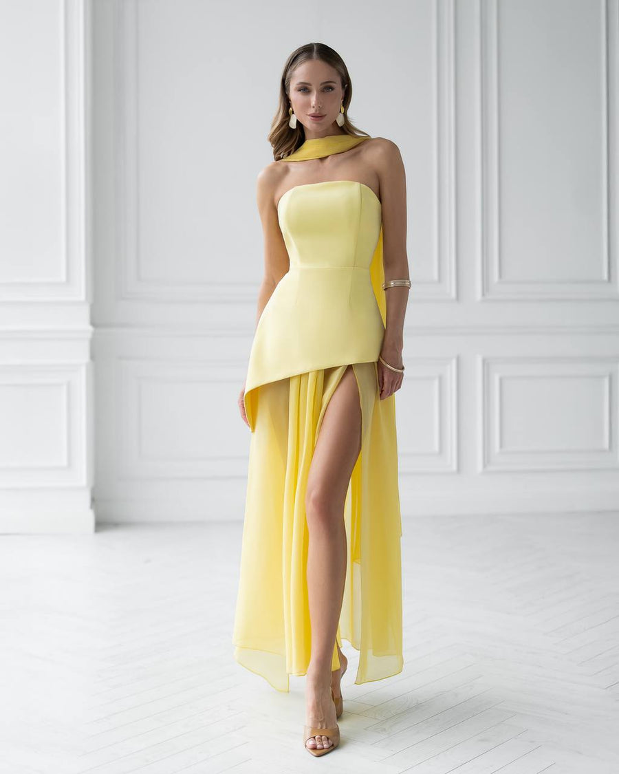 Woman wearing a yellow one-shoulder dress with a slit in a white room.