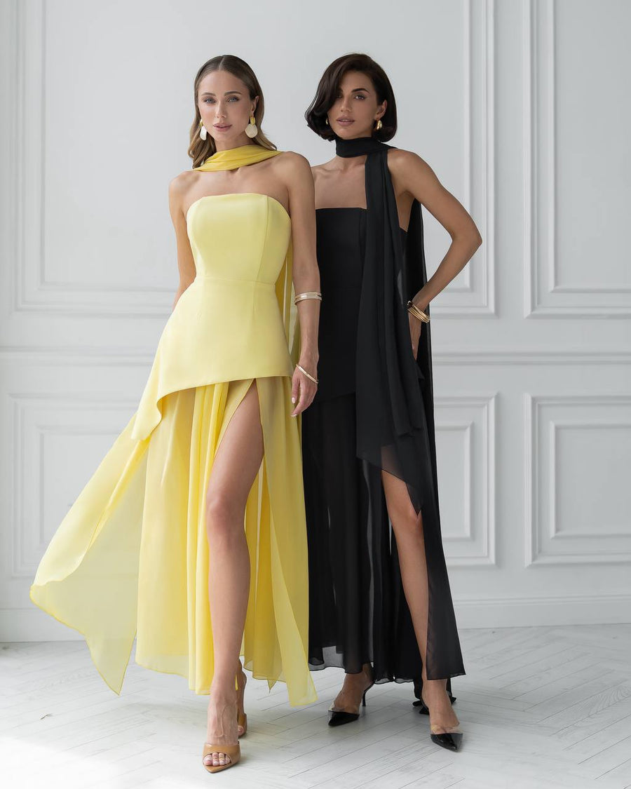 Two women wearing yellow and black evening gowns against a white wall.