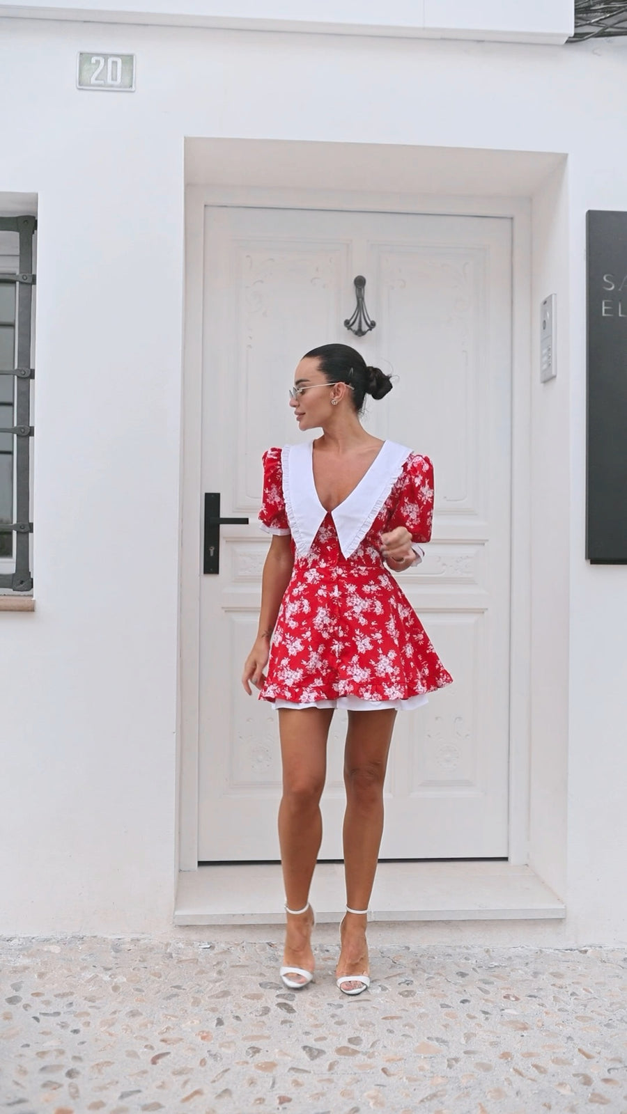 Woman wearing a red floral dress with white accents standing in front of a white door.