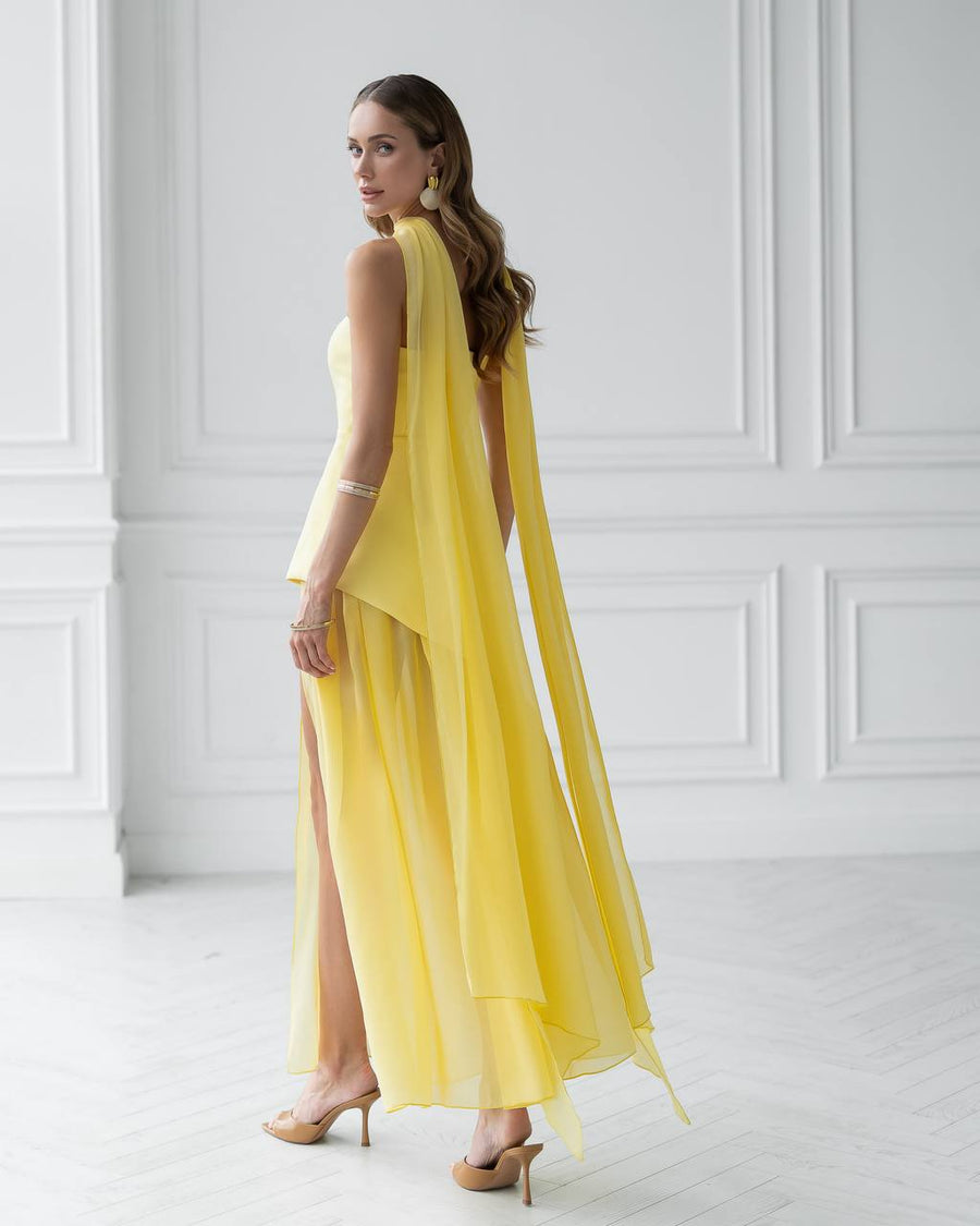 Woman wearing a yellow dress with a slit, standing against a white paneled wall.