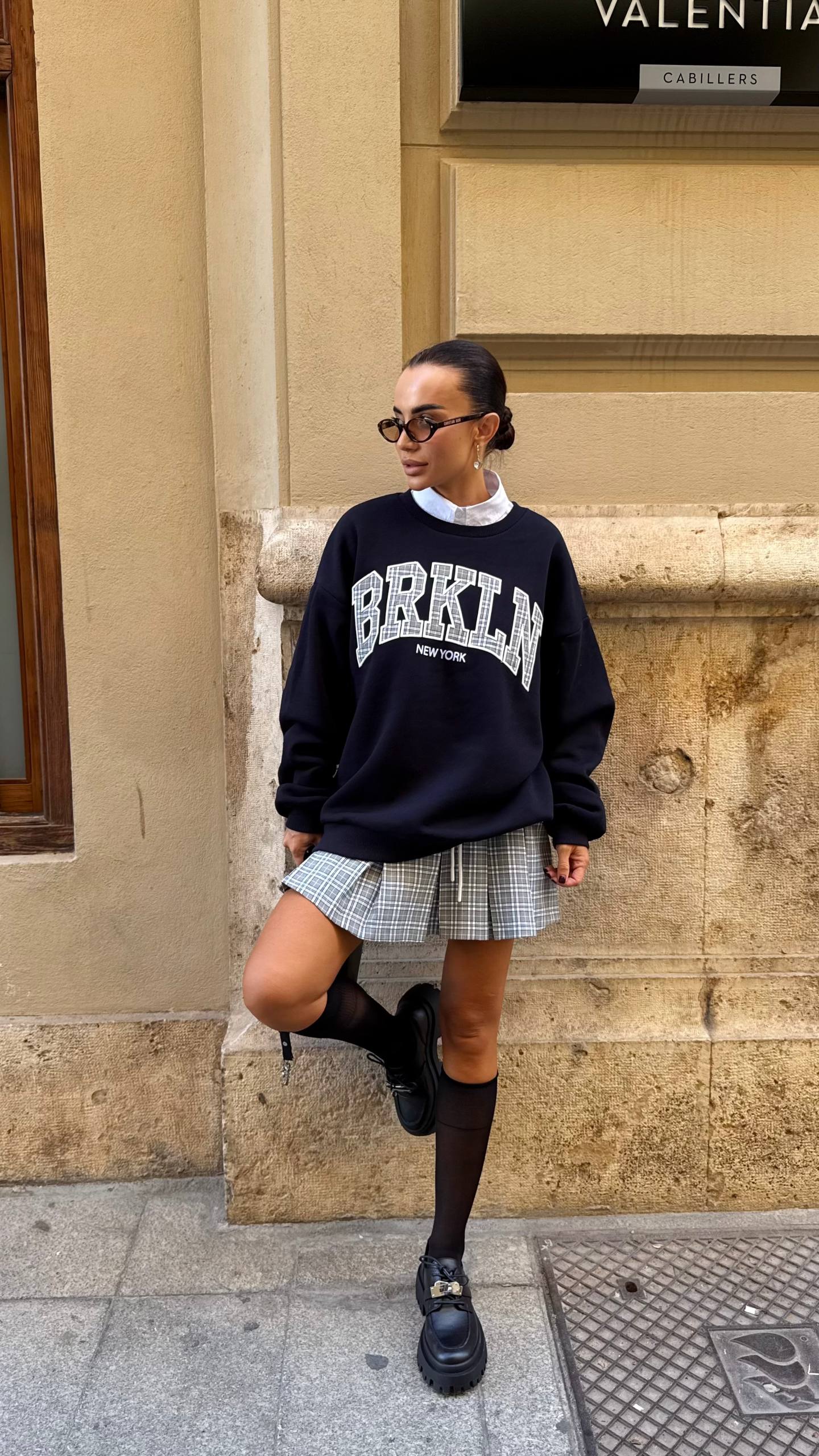 Woman wearing a black 'BRKLN' sweatshirt and plaid skirt standing against a beige wall.