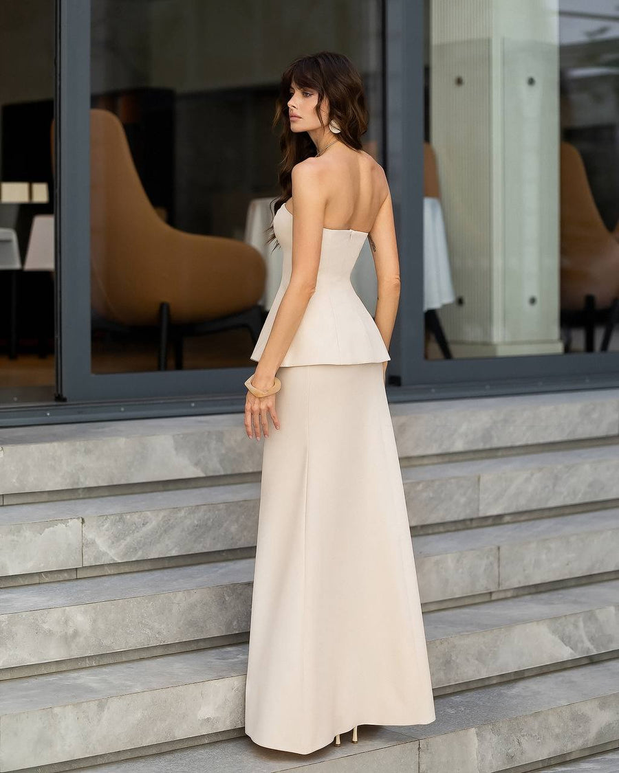A woman stands on stone steps wearing a strapless formal gown with a structured peplum waist and a long flared skirt, photographed in front of a modern building with large windows.