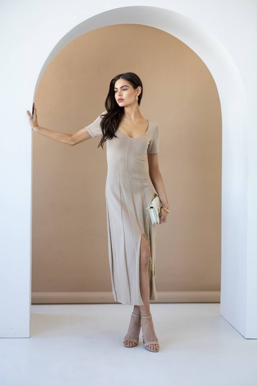 A woman models a beige short-sleeve midi dress with a square neckline and a high side slit, standing inside a white arched doorway against a warm beige background.