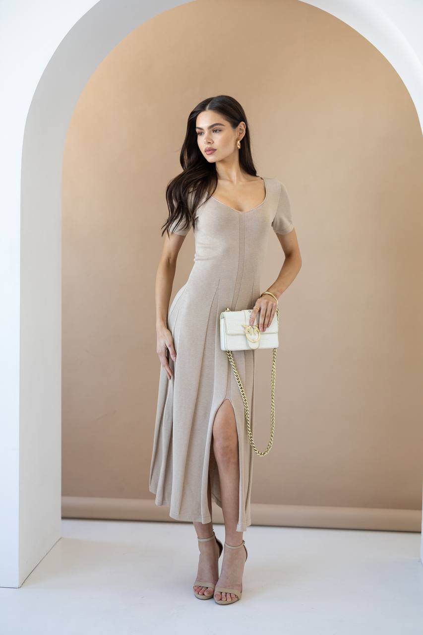 A woman models a beige short-sleeve midi dress with a thigh-high slit, holding a white handbag with a gold chain strap, set against a minimalist beige and white arched backdrop.