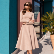 A woman wearing a long-sleeve beige A-line midi dress and black sunglasses, standing outdoors on a tiled walkway next to a teal wall and a potted palm.