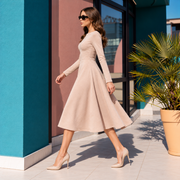 A woman wearing a light beige long-sleeve midi dress and matching nude heels walks outdoors beside a teal wall and potted palm.