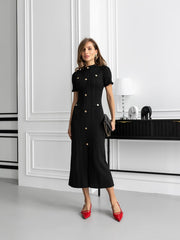 Woman in a black dress with gold buttons standing in a modern interior setting.