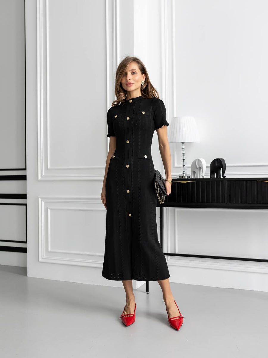 Woman in a black dress with gold buttons standing in a modern interior setting.