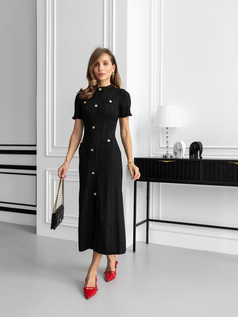 Woman in a black dress with gold buttons standing in an elegant room.