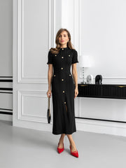 Woman in a black dress with gold buttons standing in a modern interior setting.