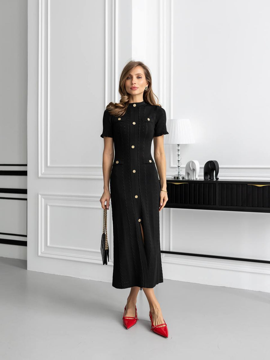 Woman in a black dress with gold buttons standing in a modern interior setting.