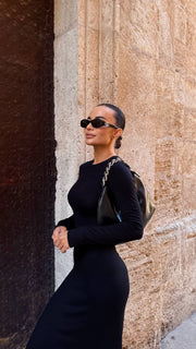 Woman in a black dress and sunglasses standing against a textured wall.