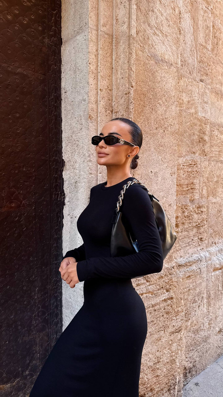 Woman in a black dress and sunglasses standing against a textured wall.