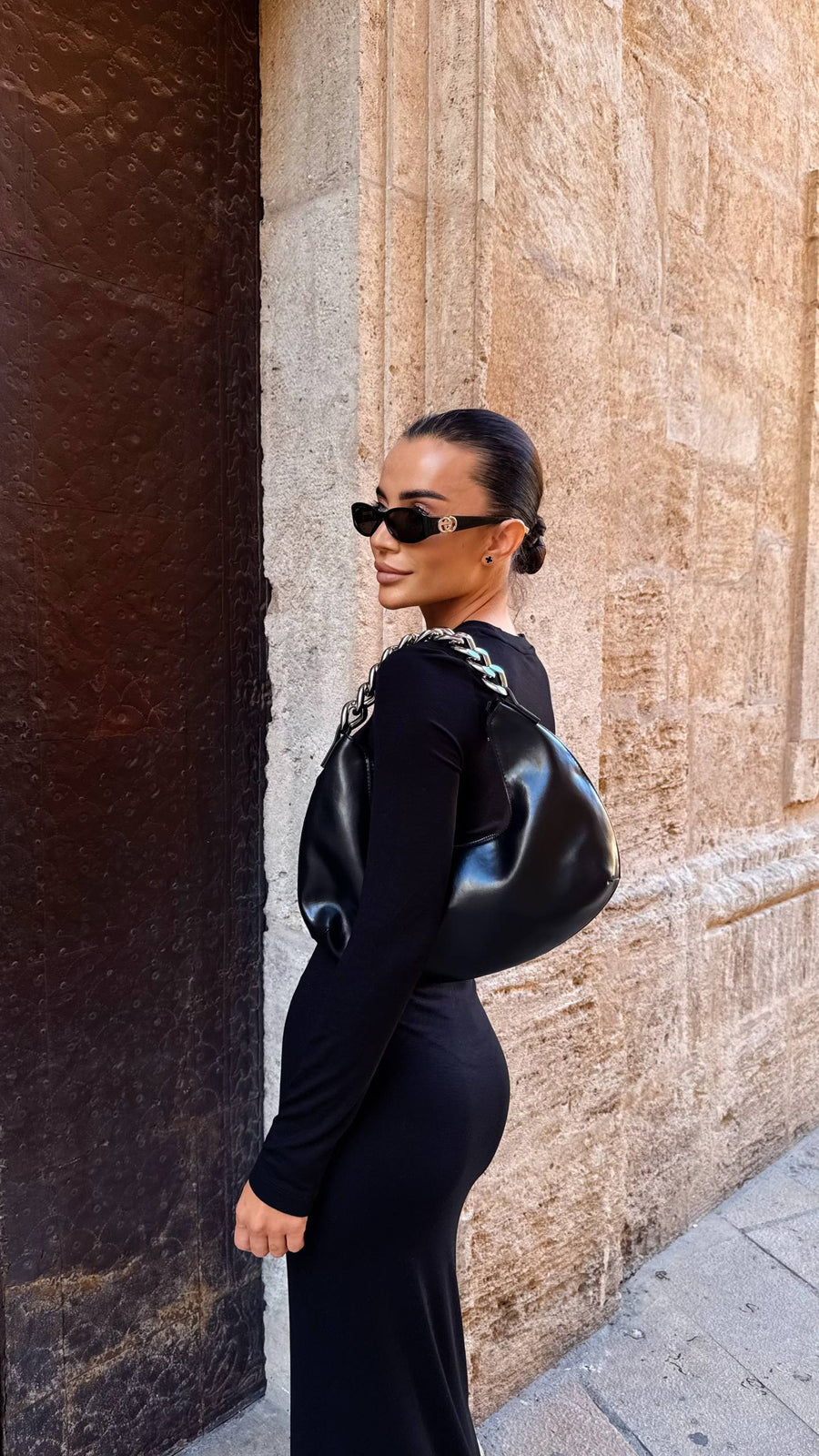 Woman in a black outfit with a shiny bag against a stone wall.