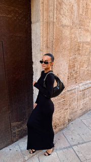 Woman in a black dress and sunglasses standing against a textured wall.