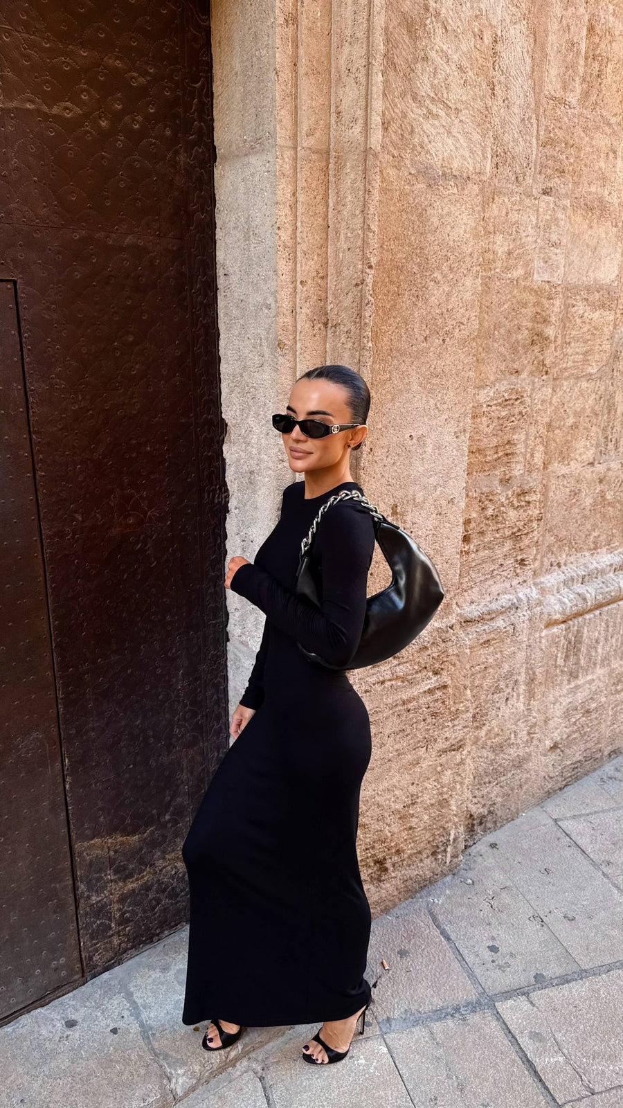 Woman in a black dress and sunglasses standing against a textured wall.