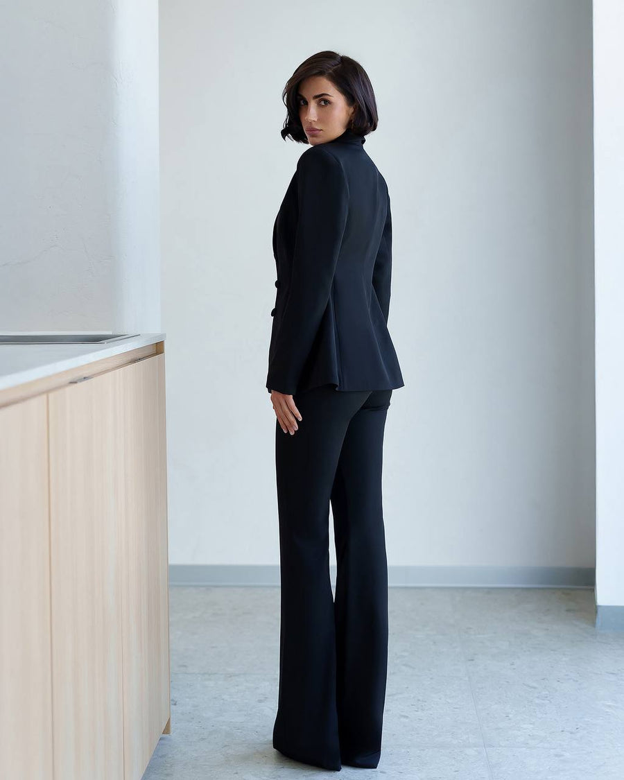 A woman stands in profile wearing a black tailored blazer and matching wide-leg trousers in a minimalist indoor setting with light gray walls and a pale wood counter on the left.