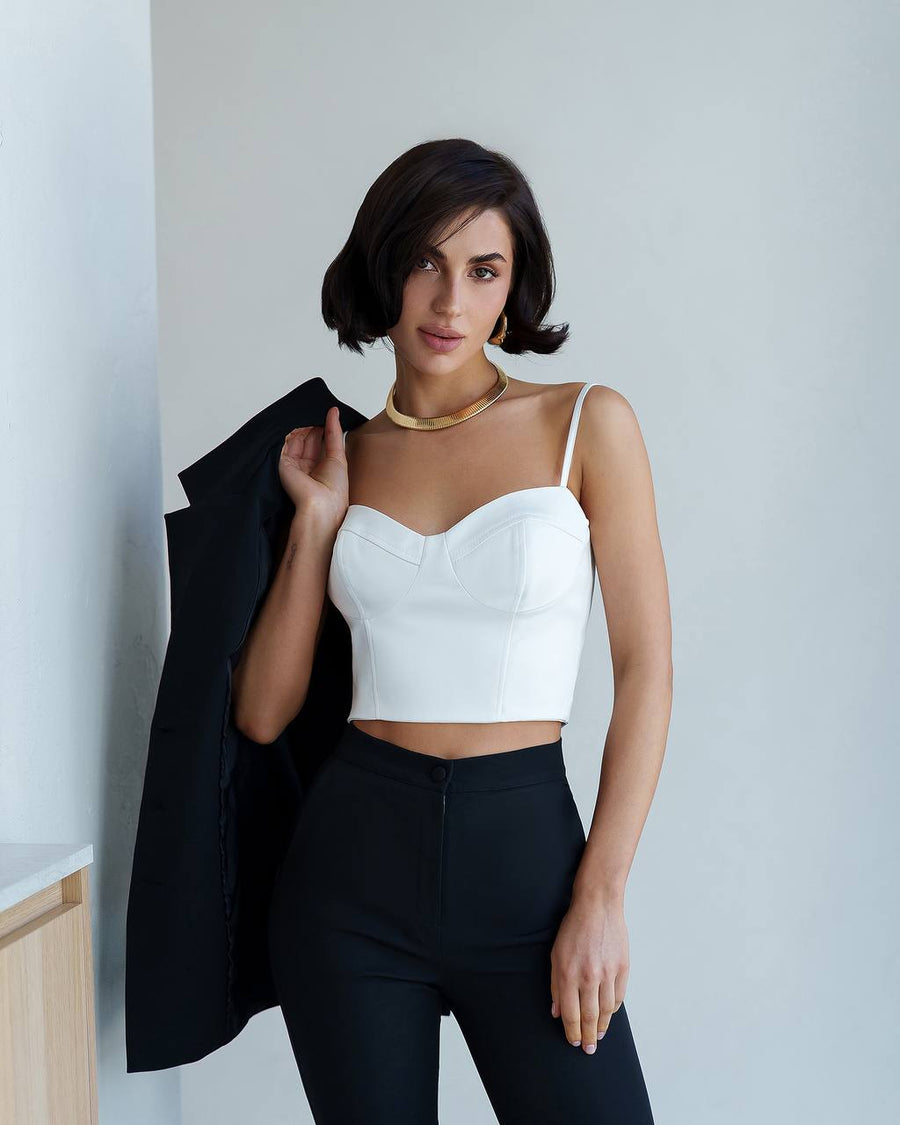 Woman wearing a white top and black pants against a light gray wall.