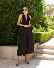 A woman wearing a black sleeveless midi dress with a plunging V-neckline and a flared skirt, standing outdoors in front of a green hedge next to stone steps.