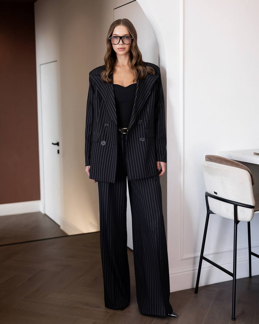 A woman wearing a black double-breasted pinstripe blazer and matching wide-leg trousers over a black top, standing in a modern indoor setting with a white wall, wooden floor, and a chair visible.