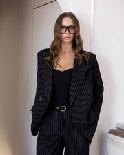 A woman wearing a black pinstripe blazer and matching trousers with a black bodysuit, styled with oversized black sunglasses, standing in a minimalist indoor setting.