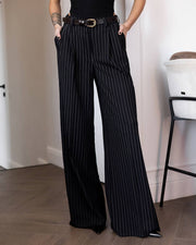 Black wide-leg pinstripe trousers with a thin black belt, modeled in an indoor setting with a wooden floor and white walls.