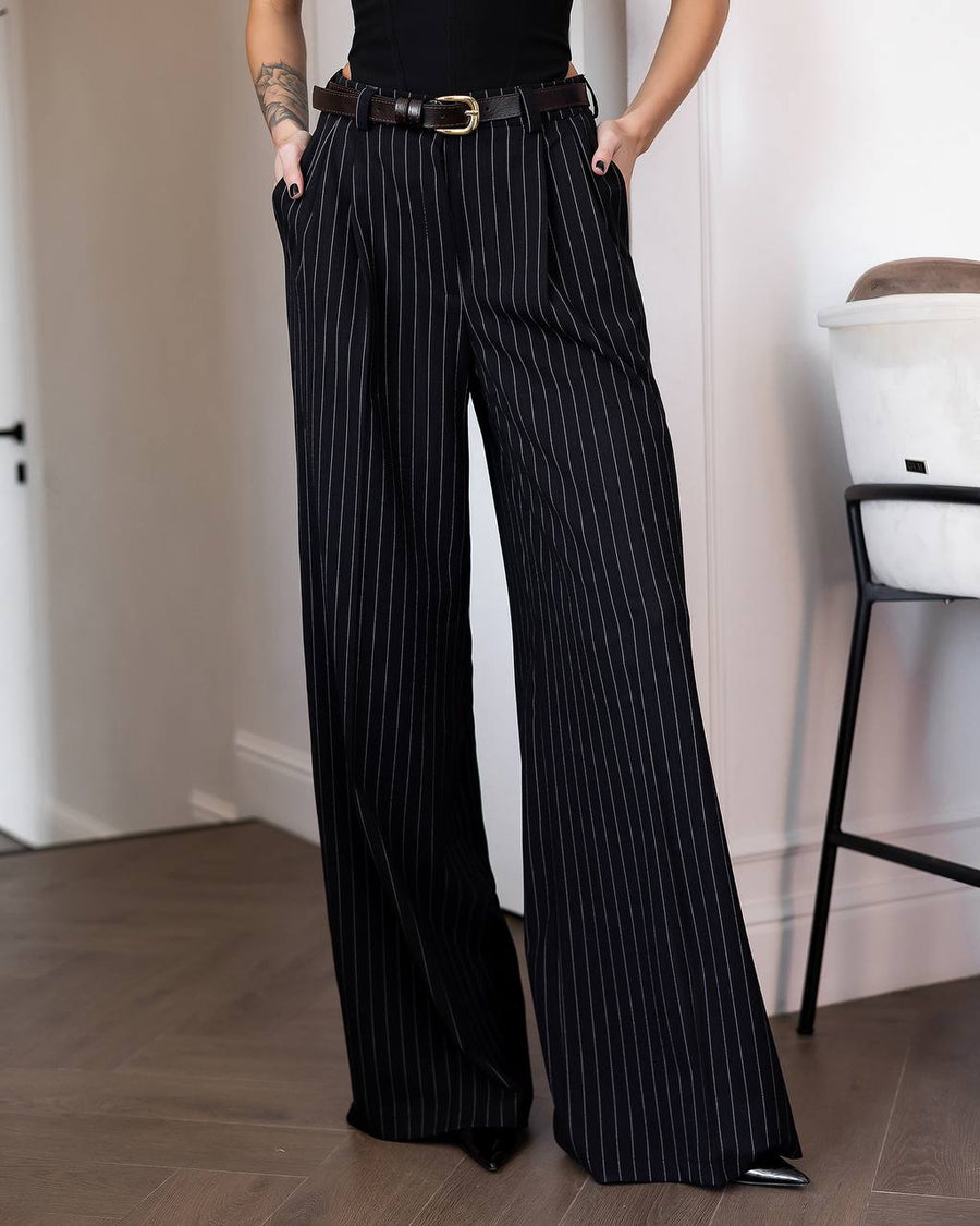 Black wide-leg pinstripe trousers with a thin black belt, modeled in an indoor setting with a wooden floor and white walls.