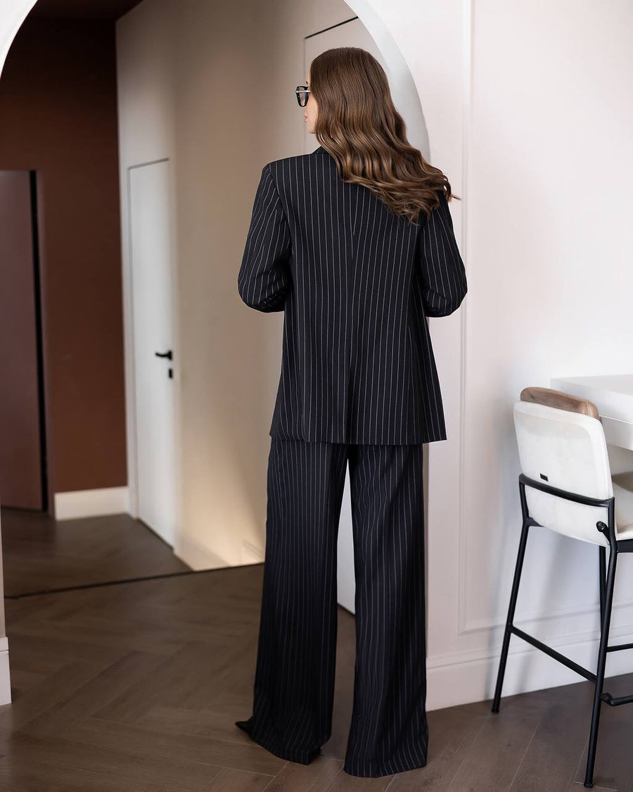 A woman stands with her back to the camera wearing a matching black pinstripe blazer and wide-leg trousers set in a modern indoor room with light-colored walls and wood flooring.