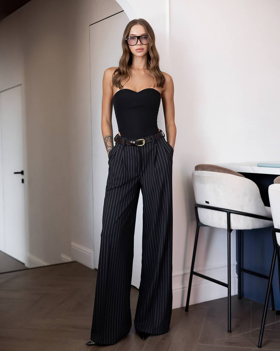 A woman stands indoors wearing a black strapless bodysuit with high-waisted black pinstripe wide-leg trousers and a slim brown belt with a gold buckle. She is wearing glasses and standing in a modern room with light walls, wood flooring, and white chairs.