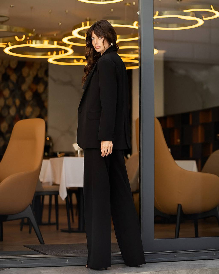 A woman stands in a modern restaurant or cafe doorway wearing a black tailored blazer and matching wide-leg trousers. The setting includes warm amber lighting, tan chairs, and glass reflections.