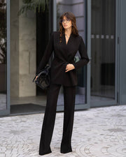A woman stands outdoors in front of a modern glass building entrance wearing a tailored black blazer and matching wide-leg trousers, holding a black handbag.