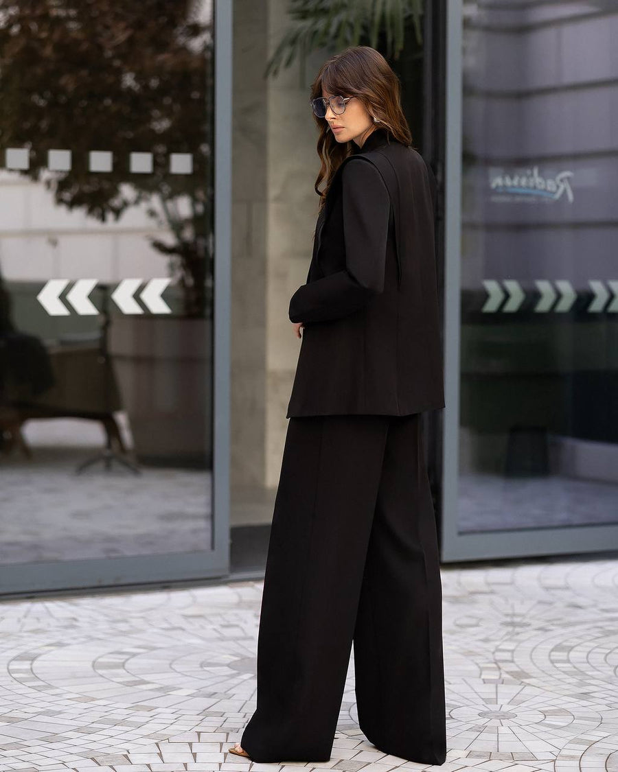 A woman wearing a black tailored suit with a sleeveless structured jacket and wide-leg trousers, standing outdoors in front of glass storefront windows.