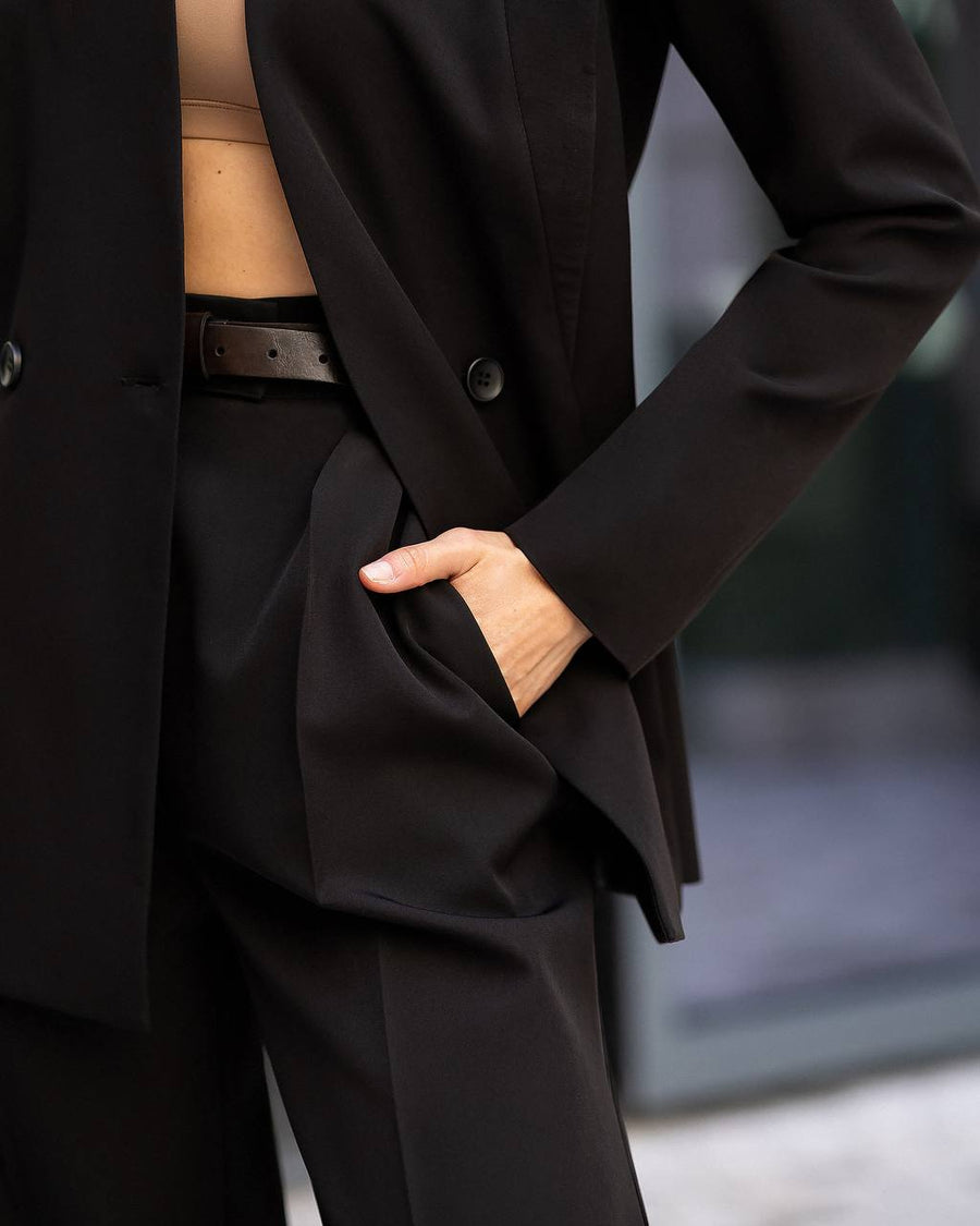 A close-up of a person wearing a tailored black blazer and matching trousers with a dark belt, with one hand tucked into the trouser pocket.