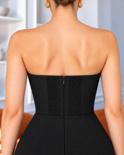 Rear view of a woman wearing a strapless black dress with a structured corset-style back panel and center zipper, shown indoors against a softly blurred light background.
