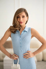 A woman modeling a light blue sleeveless polo-knit dress with a button-front collar, standing indoors with hands on hips and holding a small white handbag.