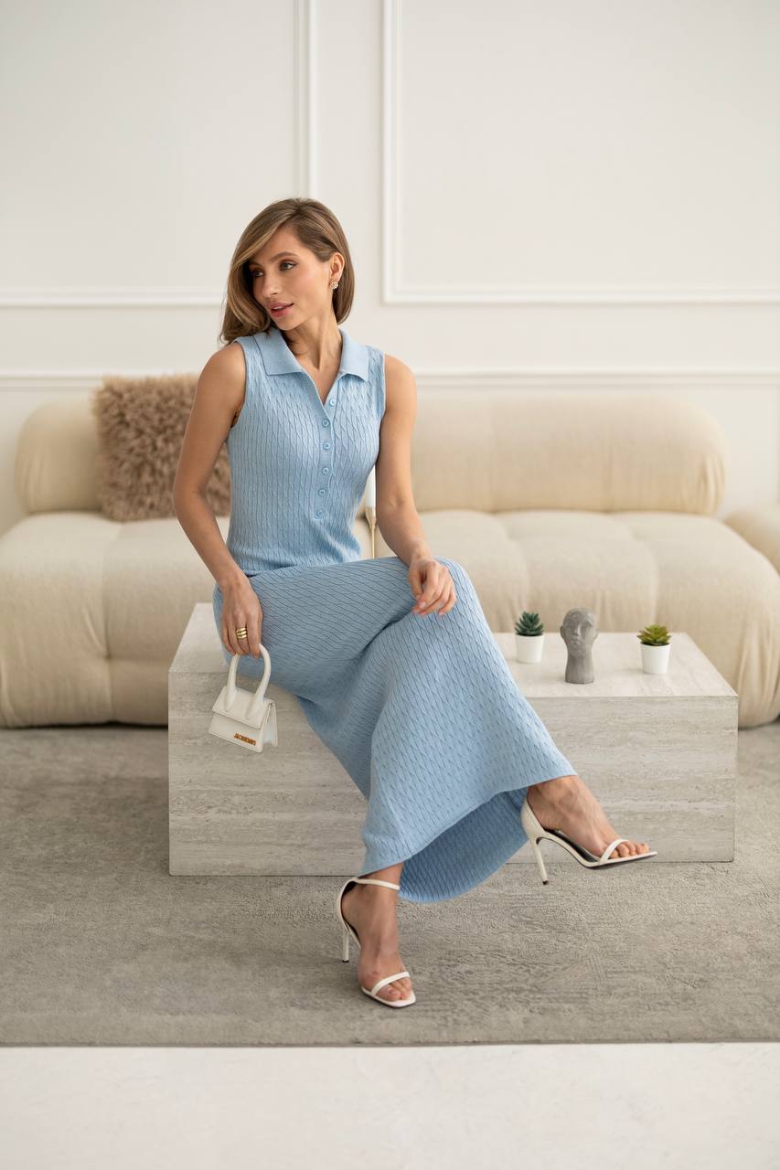 A woman models a light blue ribbed knit sleeveless collared dress with a matching skirt, sitting on a modern stone coffee table in a bright, minimalist living room.