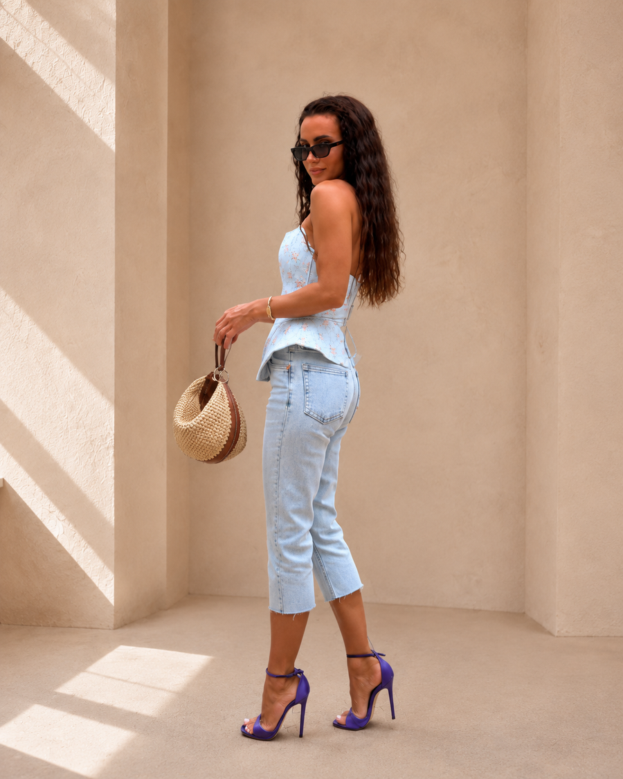 A woman stands in a sunlit beige architectural setting wearing a light blue sleeveless top and light blue cropped jeans, with purple high heels and a woven straw bag.