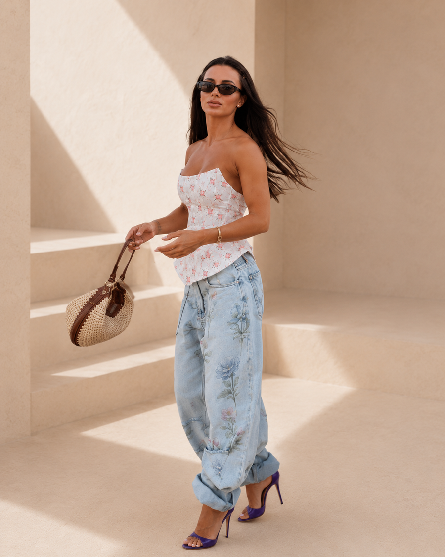A woman models a white strapless tube top with a delicate pink floral print, paired with light-wash blue floral denim jeans, purple high heels, and a woven straw handbag, set against a minimalist beige architectural background with sharp shadows.
