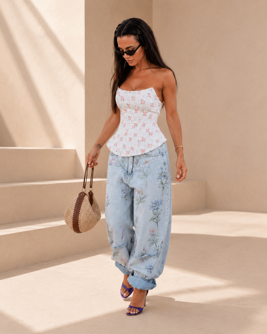 A woman walking outdoors in a sunlit architectural setting, wearing a strapless white top with a pink flamingo print, light blue floral wide-leg jeans, and purple heeled sandals, while carrying a woven straw bag.