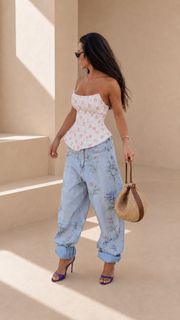A woman stands in a sunlit beige architectural setting wearing a white strapless top with a pink floral print, light blue floral jeans, purple high heels, and holding a woven straw handbag.