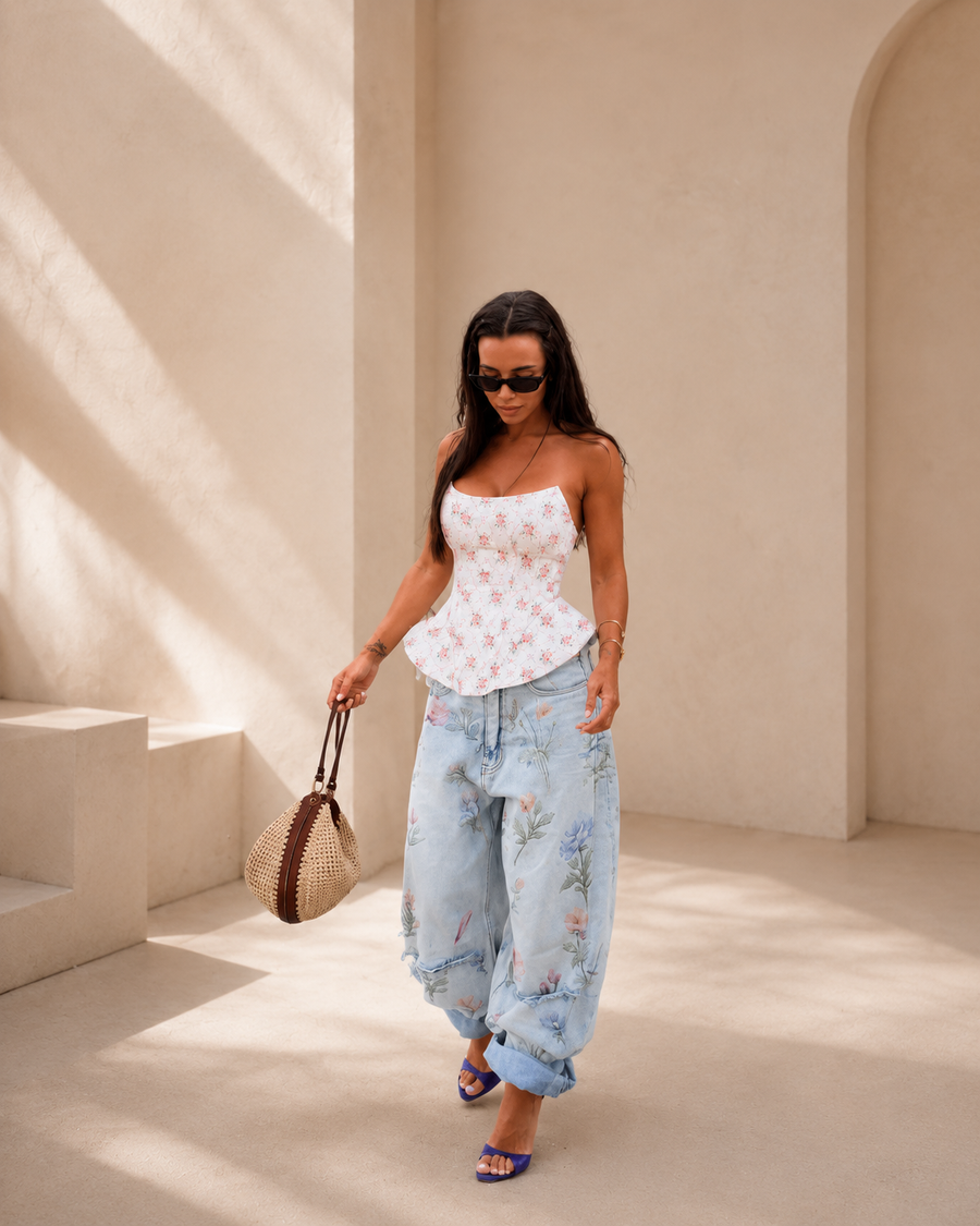 A woman walking in a sunlit architectural setting wearing a white strapless top with a pink floral print, light blue distressed jeans with a floral pattern, and blue sandals, while carrying a woven straw bag.