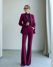 A woman modeling a matching magenta-purple tailored pantsuit with a structured blazer and wide-leg trousers, worn over a black top in a minimalist indoor setting.