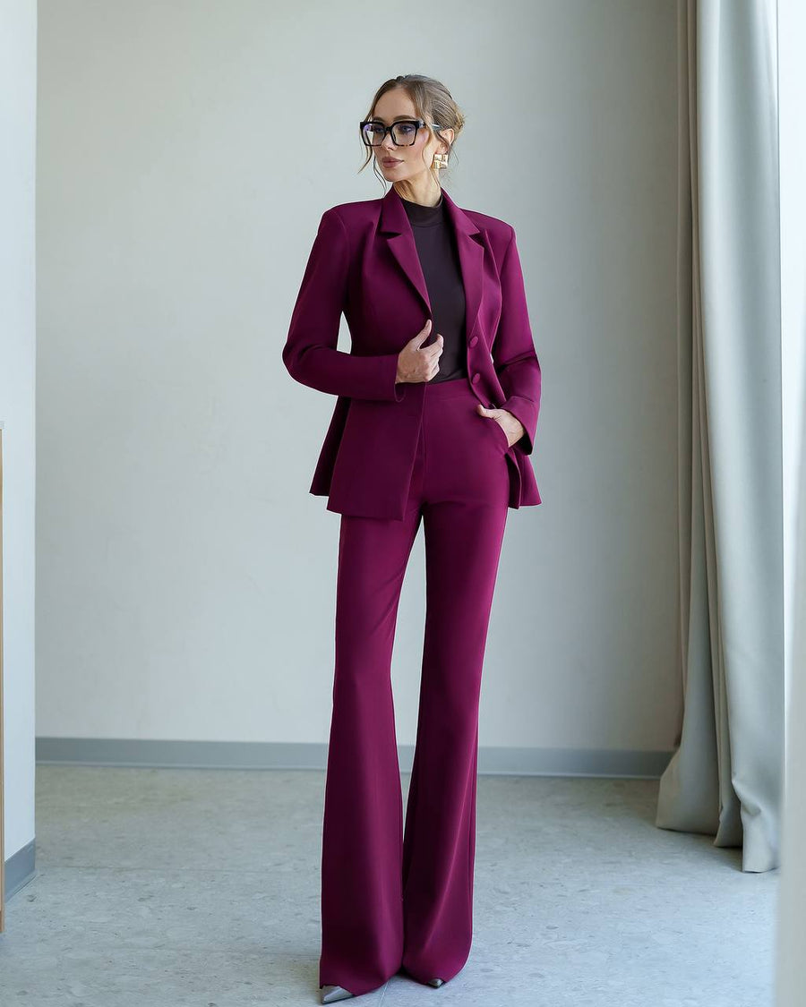 A woman modeling a matching magenta-purple tailored pantsuit with a structured blazer and wide-leg trousers, worn over a black top in a minimalist indoor setting.