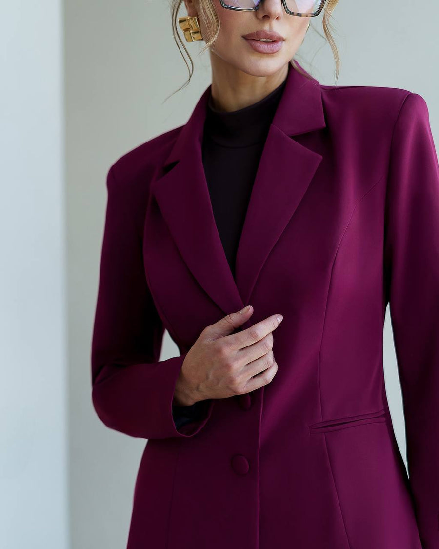 A woman wearing a structured plum-purple blazer over a black turtleneck, shown from the nose down against a plain light gray background.