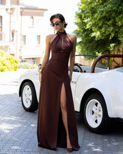 Woman in a brown evening gown standing next to a vintage car on a street.