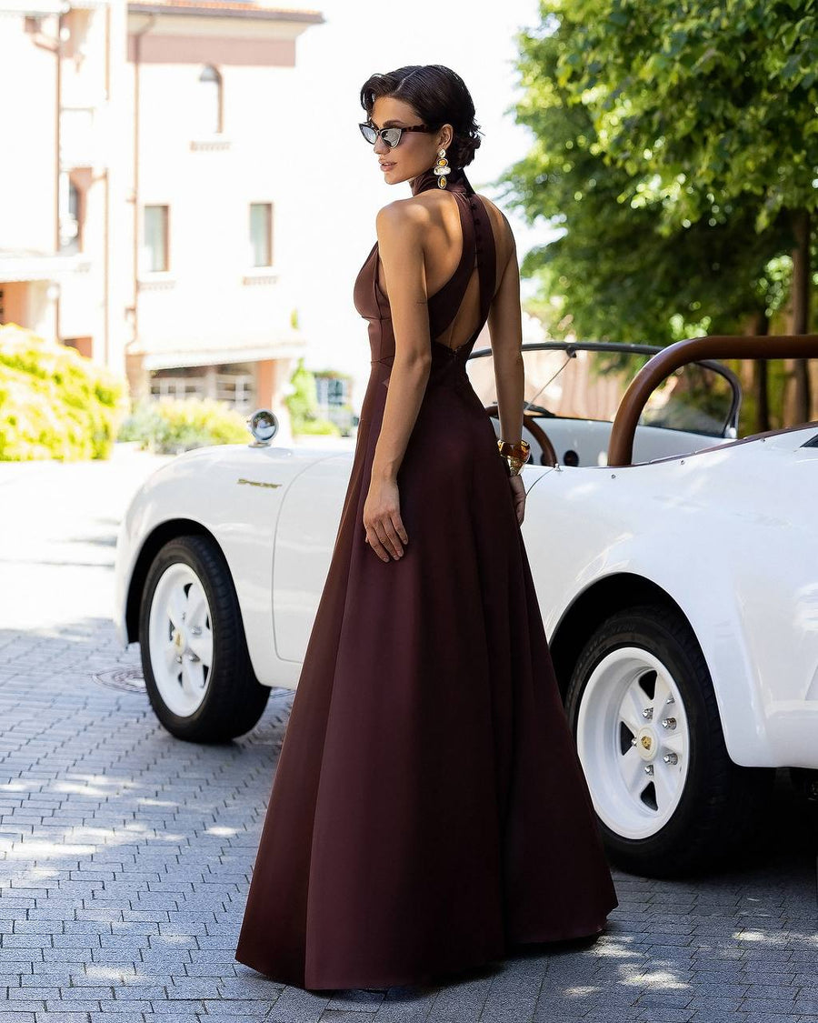 Woman in a brown evening gown standing next to a white vintage car on a sunny day.