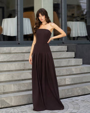 A woman models a strapless dark brown maxi dress with a fitted bodice and flowing skirt, standing on stone steps in front of a modern building with large windows.