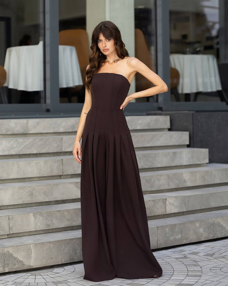 A woman models a strapless dark brown maxi dress with a fitted bodice and flowing skirt, standing on stone steps in front of a modern building with large windows.