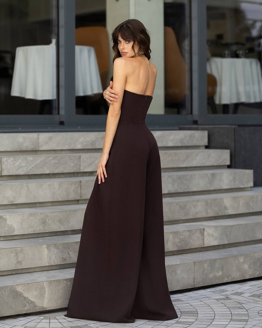 A woman wearing a strapless, floor-length dark brown gown, photographed from behind as she stands on stone steps in front of a modern building with large glass windows.