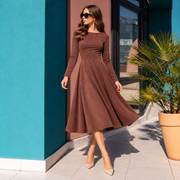 A woman wearing a brown long-sleeve midi dress with a flared skirt, styled with beige heels and sunglasses, standing outdoors next to a teal wall and a potted palm.